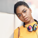 A person wearing a yellow shirt and colorful headphones around their neck leans against a wall.